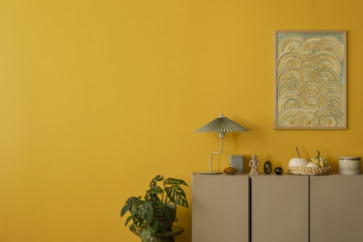 A minimalist room with a mustard yellow wall, abstract art, a lamp, plants, and decorative items on a beige cabinet.