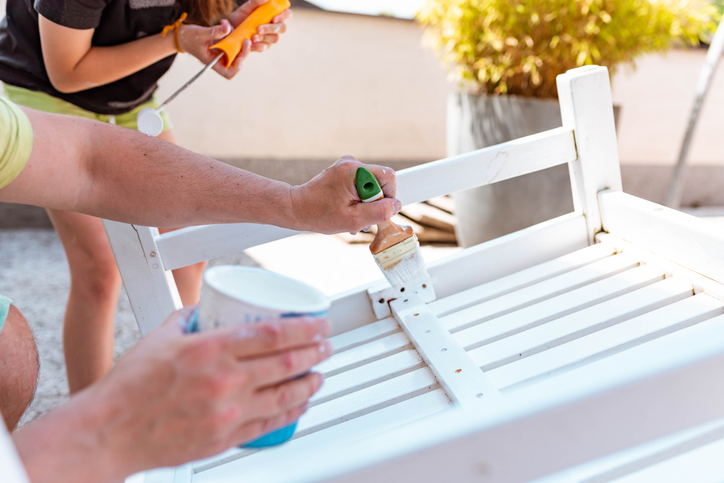 Two people painting a white wooden chair outdoors.
