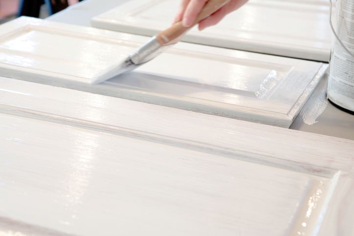 A hand painting wooden cabinet doors white with a brush, next to a can of paint on a table.