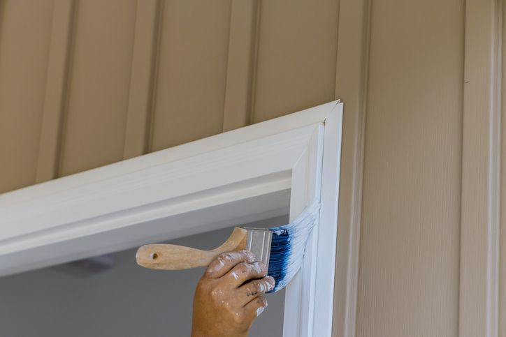 Hand painting a white door frame with a brush against a beige paneled wall.