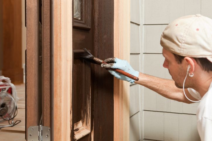 Person painting a wooden door with a brush, wearing a cap, gloves, and earbuds, focusing on applying brown paint to the door panel.