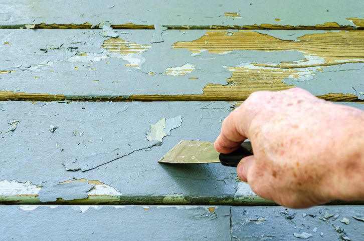 A hand scraping peeling blue paint off wooden planks with a putty knife, revealing the natural wood underneath.