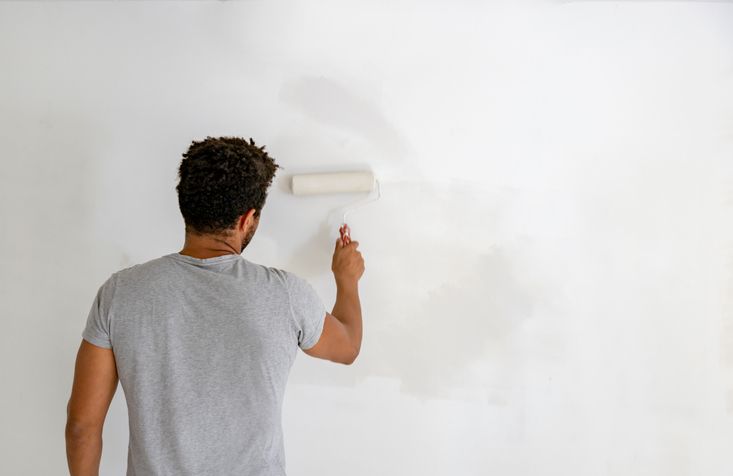 Person painting a white wall with a roller, wearing a grey t-shirt. The wall is patchy.
