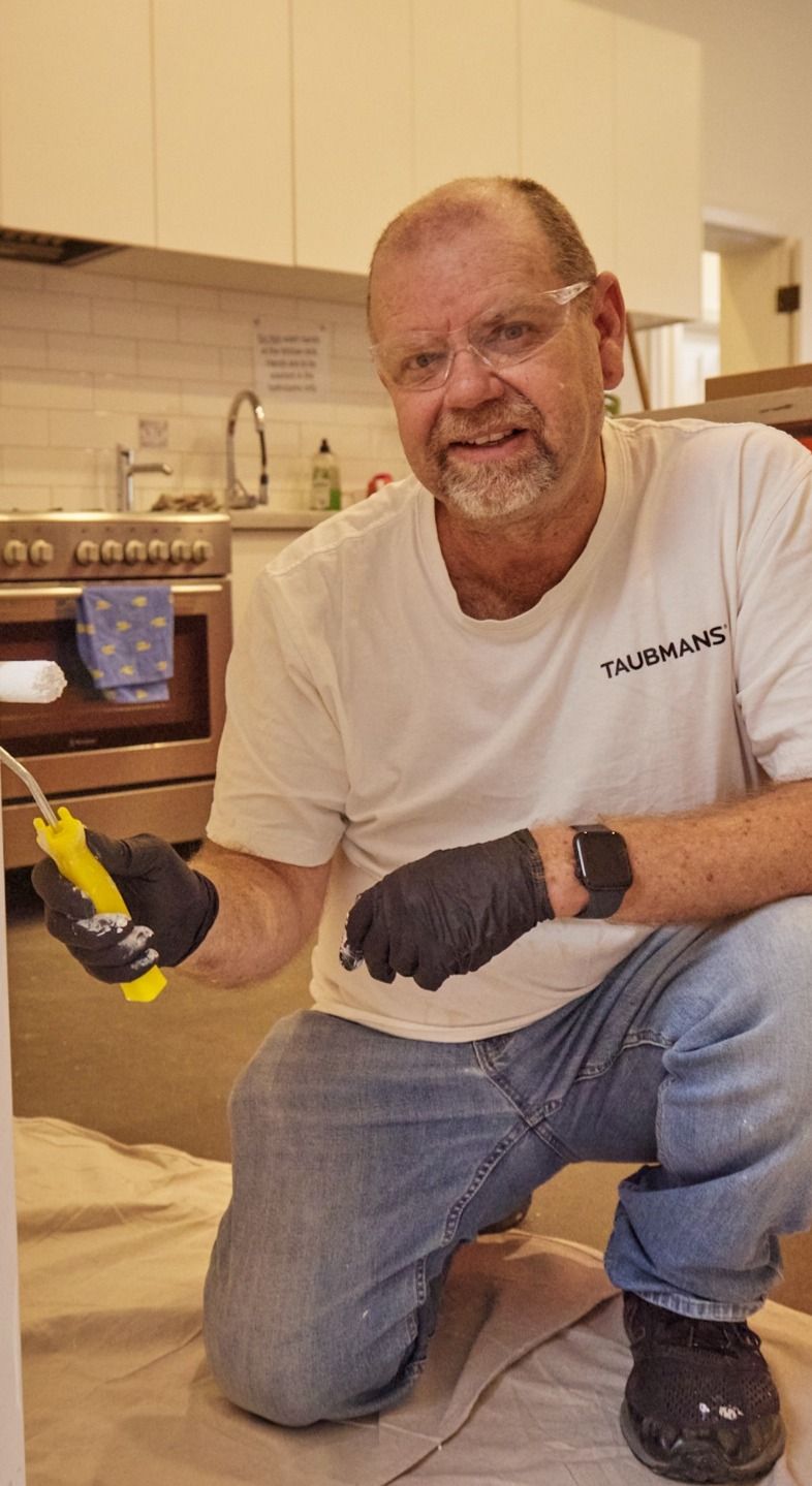 A man in a white shirt and gloves is painting a wall with a small roller in a kitchen. He is smiling and wearing safety glasses.