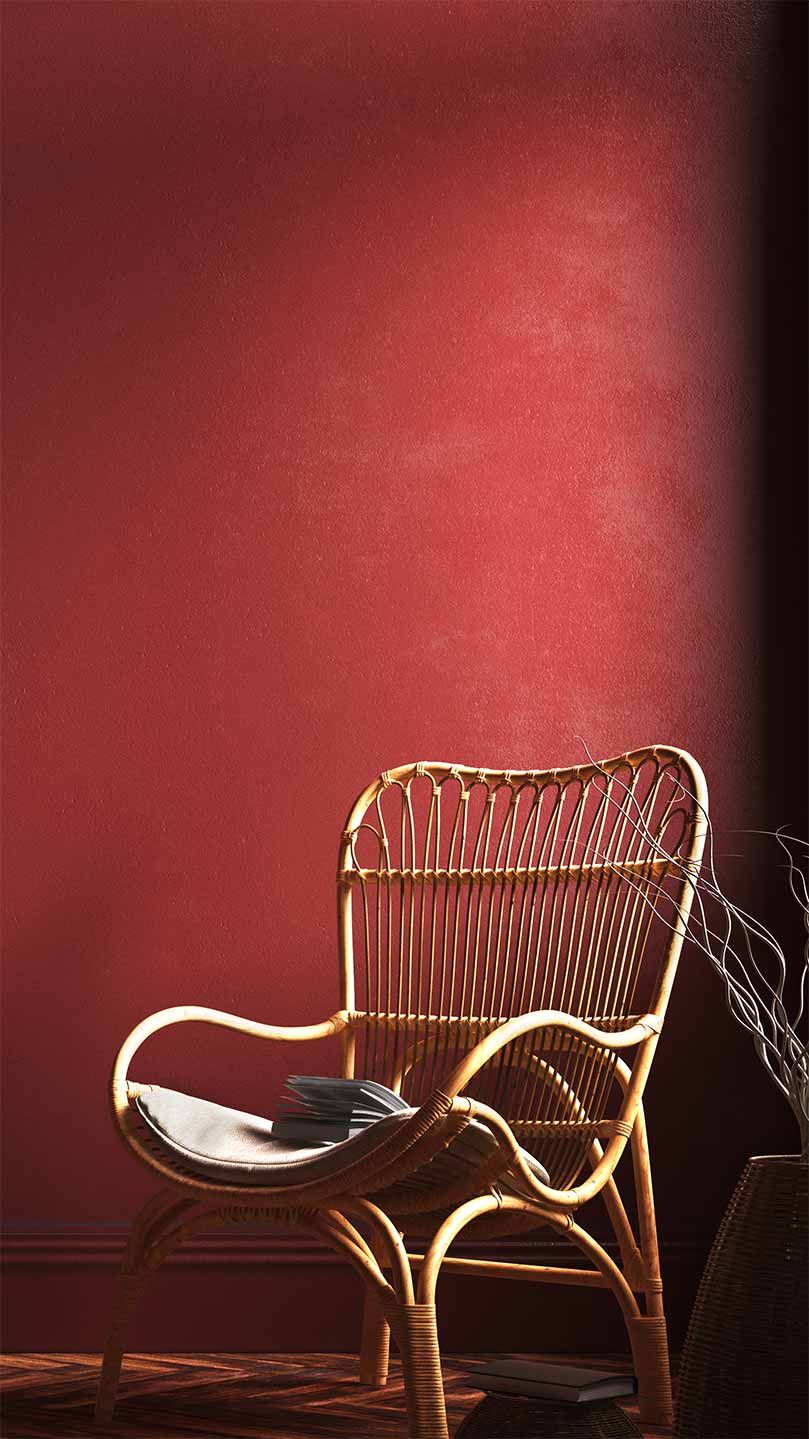 Wicker chair with a cushion and book on a wooden herringbone floor against a deep red wall, next to a vase with twigs.