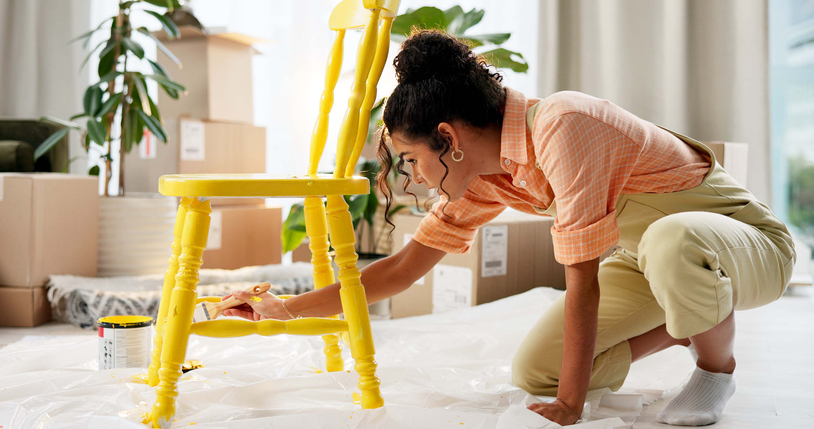 Person painting a wooden chair yellow in a room with plants and boxes, kneeling on a plastic-covered floor.