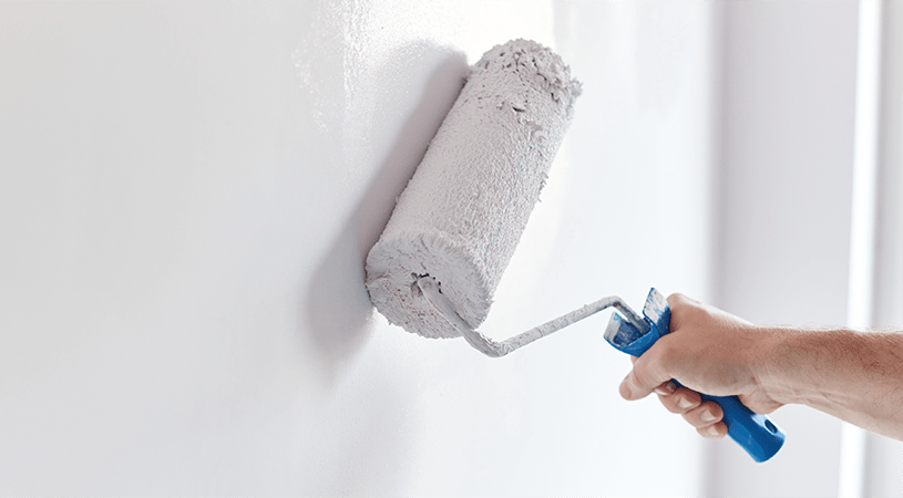 A hand using a blue-handled paint roller to apply white paint on a smooth wall.