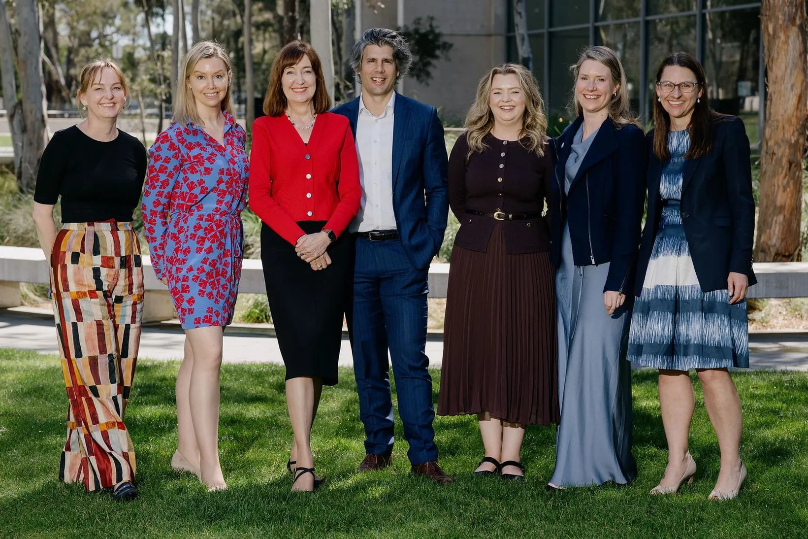 A group of six women and one man stand smiling on a grassy area in front of a modern glass building. They are dressed in smart, colorful outfits.