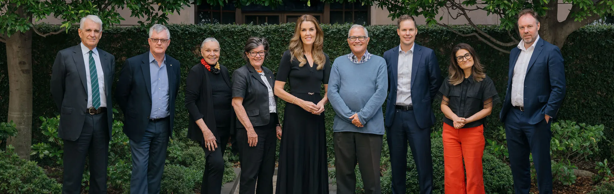 A group of nine people, dressed in business attire, stand in a row by a fountain, with trees and greenery in the background.