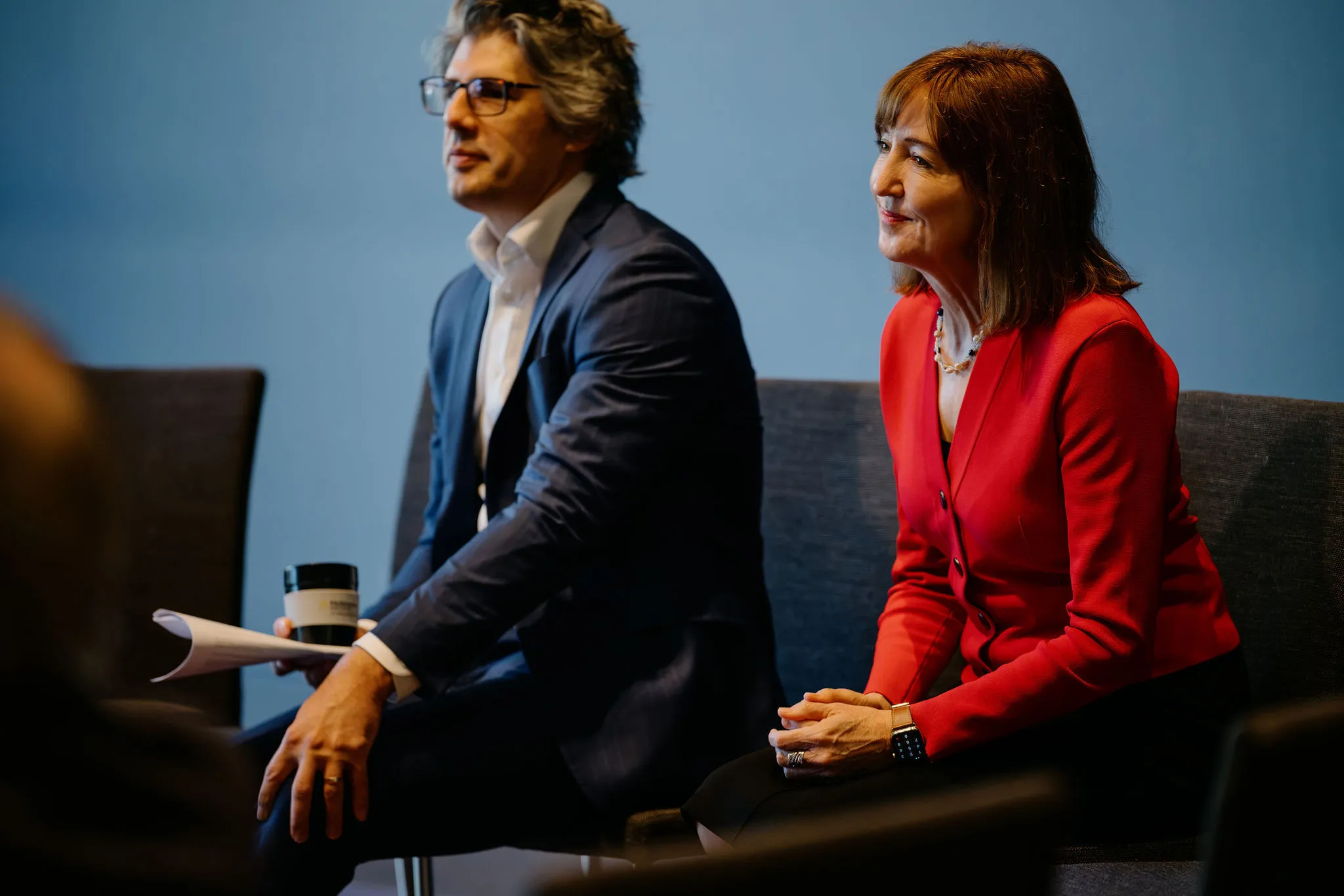 A man in a blue suit and a woman in a red jacket sit attentively, with the man holding a paper and a cup.
