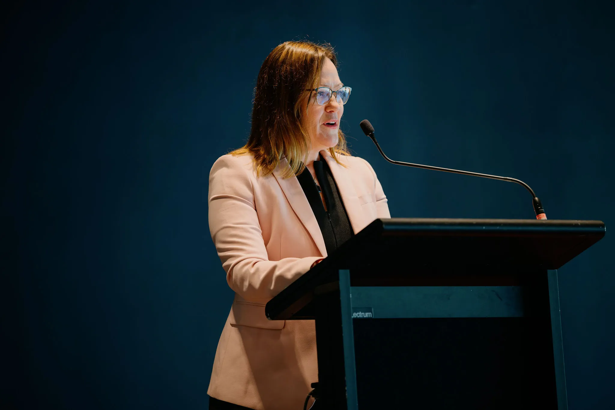 A woman in a pink blazer speaks into a microphone at a podium against a dark blue background.