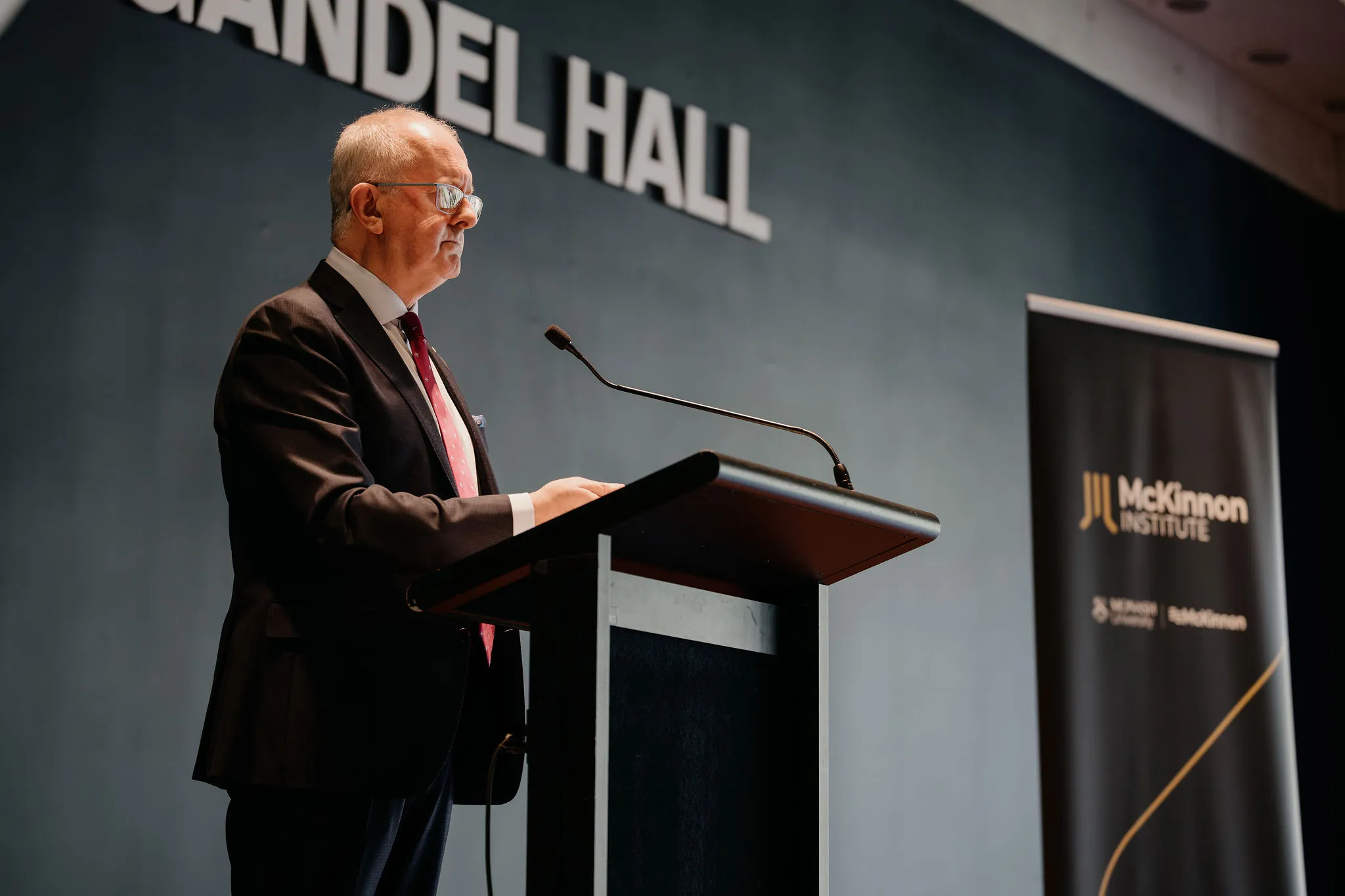 A man in a suit speaks at a podium in Gandel Hall, with a banner for the McKinnon Institute in the background.