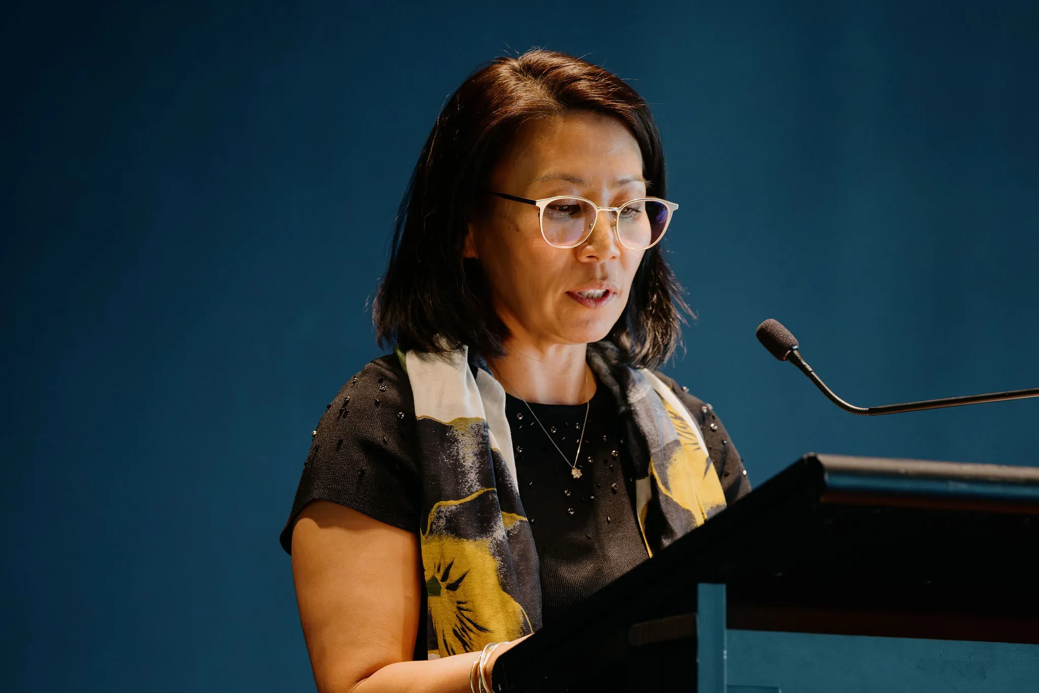 A woman with glasses and a floral scarf speaks at a podium, set against a blue background.