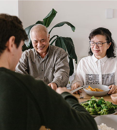 Three people sitting at a dining table, smiling and serving food. The table has bowls of broccoli and rice. A large plant is in the background.