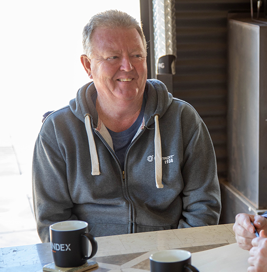 Smiling man in a gray hoodie sits at a table with coffee mugs, engaged in conversation.