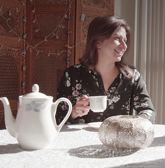Woman sitting at a table looking to the right, holding a cup of tea. Tea pot and candle holder are on the table.