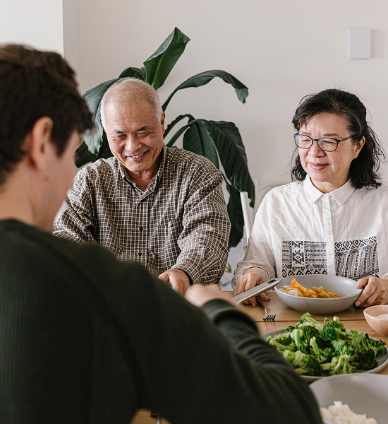 Three people enjoying a meal together at a table with bowls of food and a leafy plant in the background.