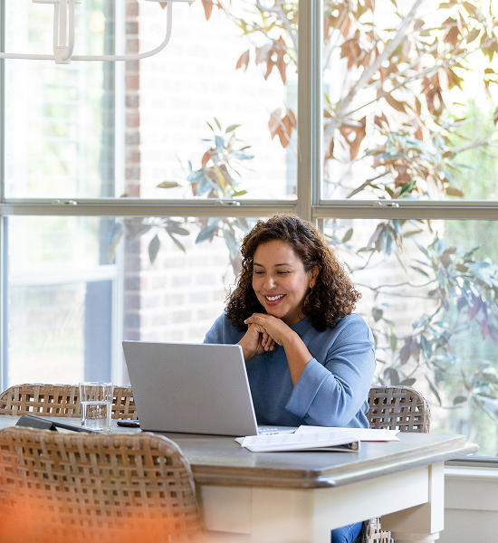 Woman smiling at a laptop in a bright room with large windows, seated at a table with papers and a glass of water.