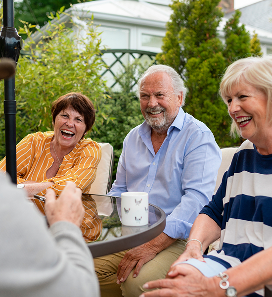 Three older adults sitting outdoors, laughing and chatting around a table with a mug, surrounded by greenery.