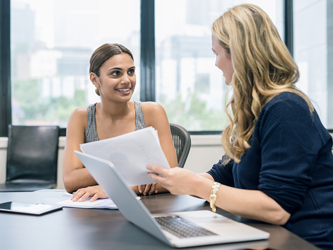 Two women sitting at a table in an office, discussing documents. One holds papers, and a laptop is open in front of them.