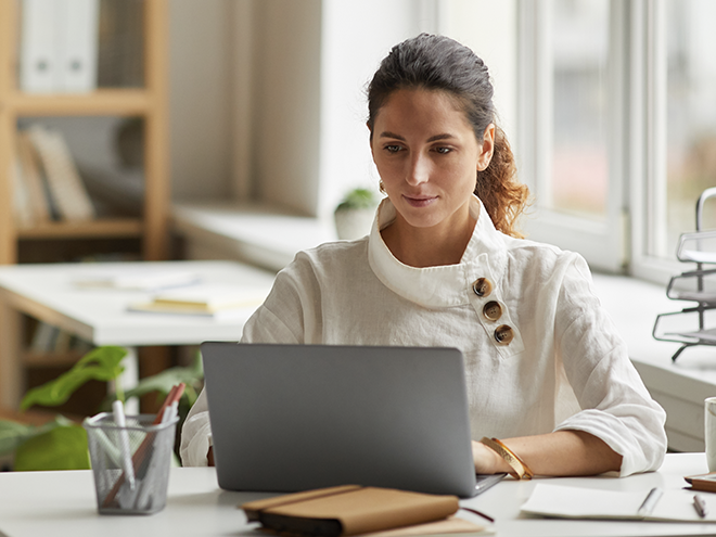 Woman in a white blouse working on a laptop at a desk, with notebooks and office supplies nearby, in a bright, modern office setting.