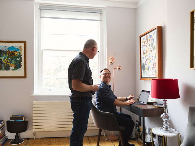 Two men in a bright room, one standing and the other seated at a desk with a laptop, smiling at each other. Art and a red lamp decorate the space.