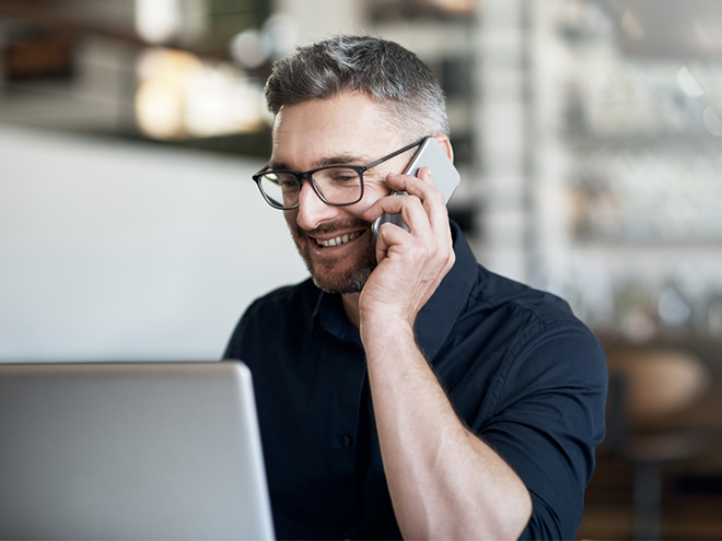 A man with glasses, smiling while talking on a smartphone, sits at a desk with an open laptop in a modern office setting.